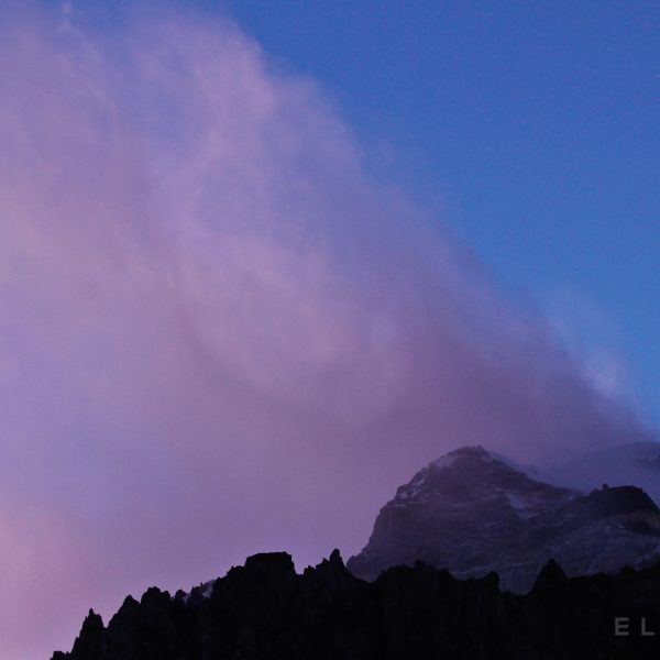 Pinksih looking clouds form around the summit of a mountain with snow leading to the summit with rocky formations below