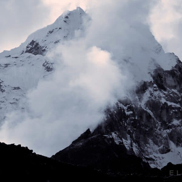 A mountain in the Himalayas stands amongst a cloudy sky with a rocky ridge in the foreground