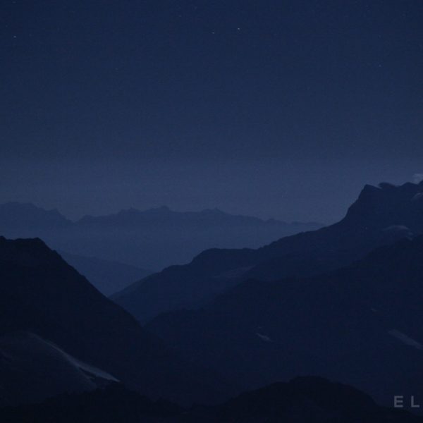 A mountain stands above a velley at night with a purpleish sky and the onset of stars