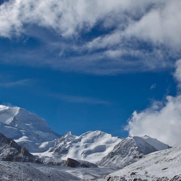 A snow capped mountain the distance surrounded with lower snow capped peaks with clouds and a blue sjy