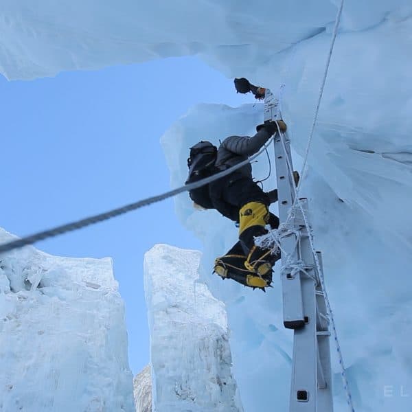 A climber climbs an aluminum ladder while wearing large yellow boots while surrounded by large untable blocks of ice