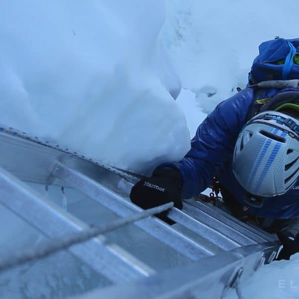 A climber dressed in blue with a blue backpack climbs an aluminum ladder  with large unstable blocks of ice