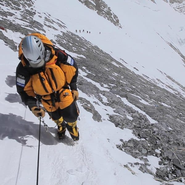 Climber dressed in orange with an oxygen mask walks with a safety line in had on snow with rocky terrain next to him with climbers in the distance