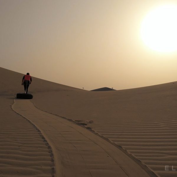 Person pulling a tire attached to a rope through the desrt dunes with some minor vegetation and a bright yellow sun