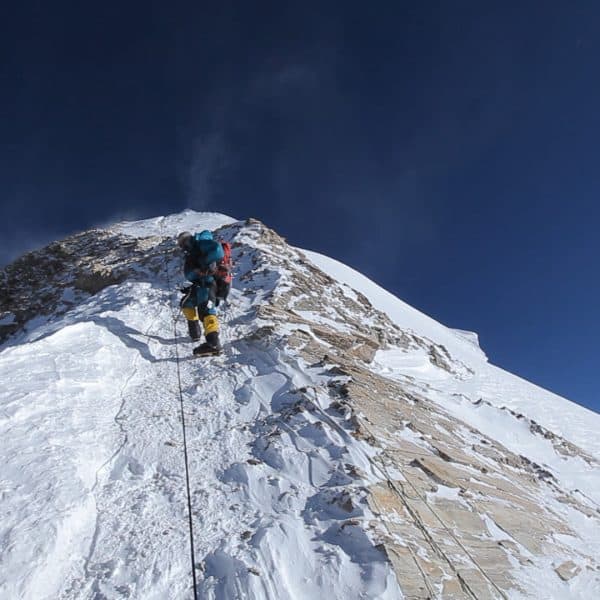 A steep narrow snowy and rocky trail near the summit of Everest with a climber breating oxygen through a mask dressed in blue