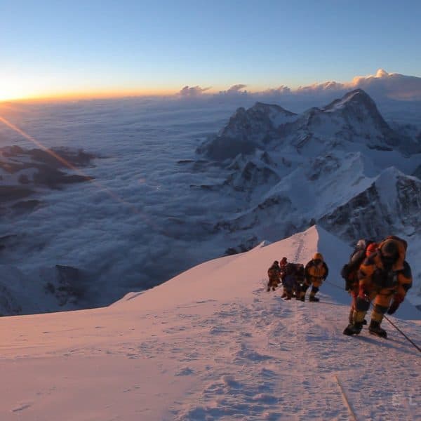 Climbers dressed in warm clothing wearing oxygen masks and orange suits on Mt Everest move slowly as the sun rises in the distance with mountains and clouds beneath them