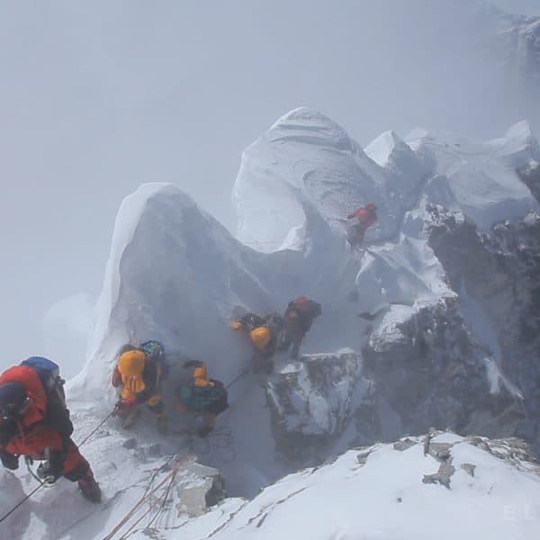 Climbers wearing multip colored downsuits approach a summti with a cornice to their right as they follow a safety rope wearing crampons as the snow blows from high winds on Mt Everest