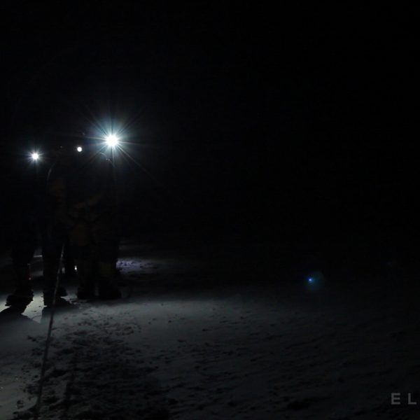 Climbers walk with headlamps at night wearing crampons on a trail of snow while holding a rope