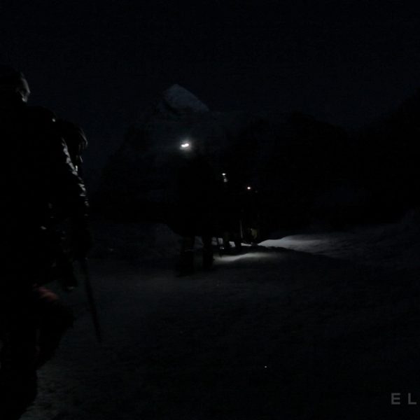 Climbers with headlamps walk in the dark with crampons and ice axes ith a faint mountain in the distance near Mt Everest