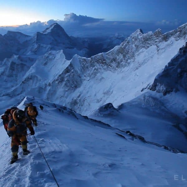 Climbers dressed in warm clothing wearing oxygen masks climb to the summit of Mt Everest with mountains and clouds beneath their feet in the distance