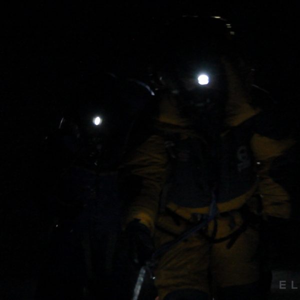 Climbers walk with headlamps at night wearing crampons on a trail of snow while holding a rope