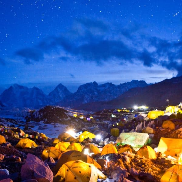 Multi colored tents lit up with headlamps on a glacier with a starry night sky with mountains in the distance