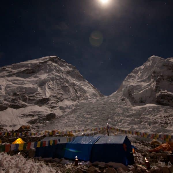 Multi colored tents at the base of a snowy glacier at night with colorful prayer flags in the Himalyas with mountains in the distance near Mt. Everest