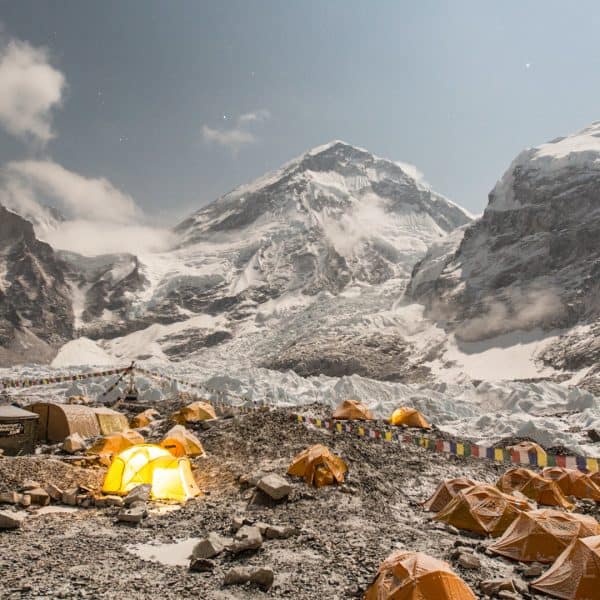 A camp on a rocky glacier at night with orange tents with clouds in the background revealing two mountain near Mt Everest
