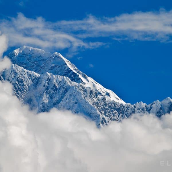 A triangular snow capped summit is surround by clouds and a vibrant blue sky
