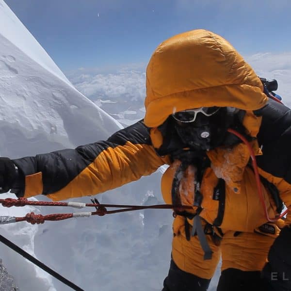 An exhausted climber breating supplemental oxygen through a mask struggles as he hangs onto a rope trying to ascend while wearing black gloves with clouds beneath him on Mt Everest