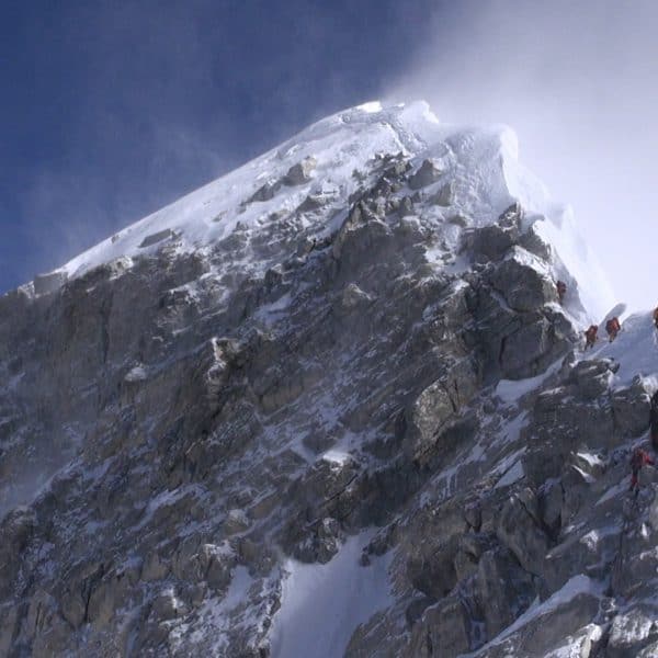 Two climbers sdressed in orange sit in the snow as other climbers climb towards them while attached to a rope with the highest mountains in the world beneath them