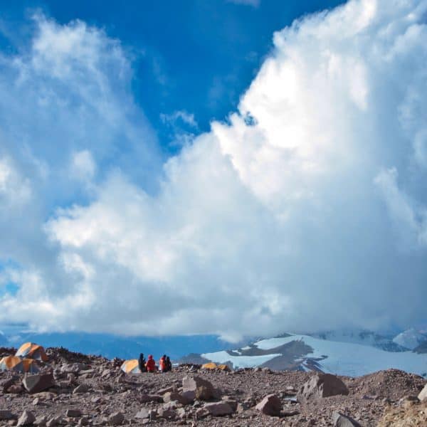 Climbers stand next to orange and grey tents on a rocky glacier with mountains and clouds in the distance