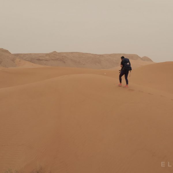 A hiker carries a backpack and walks up a desert dune with brow cliffs in the distance