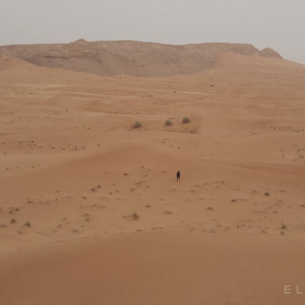 A hiker carries a backpack and walks though the sand dunes with brown rock formations in the distance