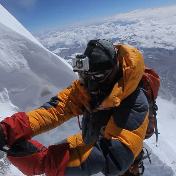 A climber breating supplemental oxygen through a mask on Mt Everest climbs a steep cliff with a rope known as the Hillary Step as other climbers dressed in blue