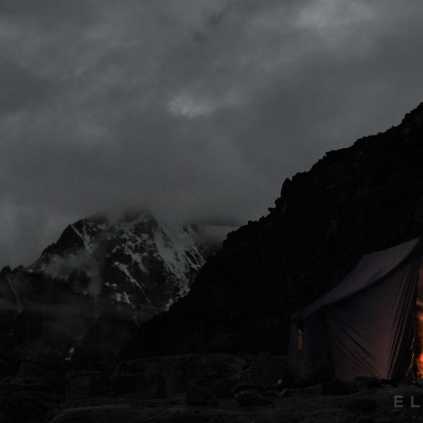 A purple tent with a light inside sits on a snowy rocky area with mountains and clouds in the background at night