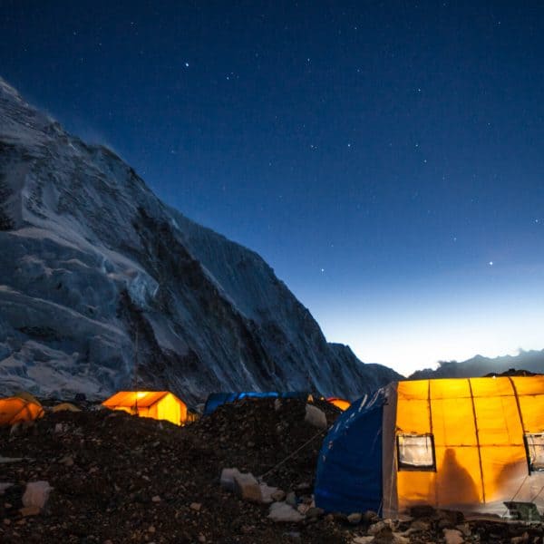 A blue tent set up on a glacier near Mt Everest with multi colored tents nearby as the sun sets revealing the stars in the distance