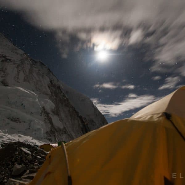 Orange tents set up on a glacier on Mt Everest as the moon sets in the distance with racing clouds at night