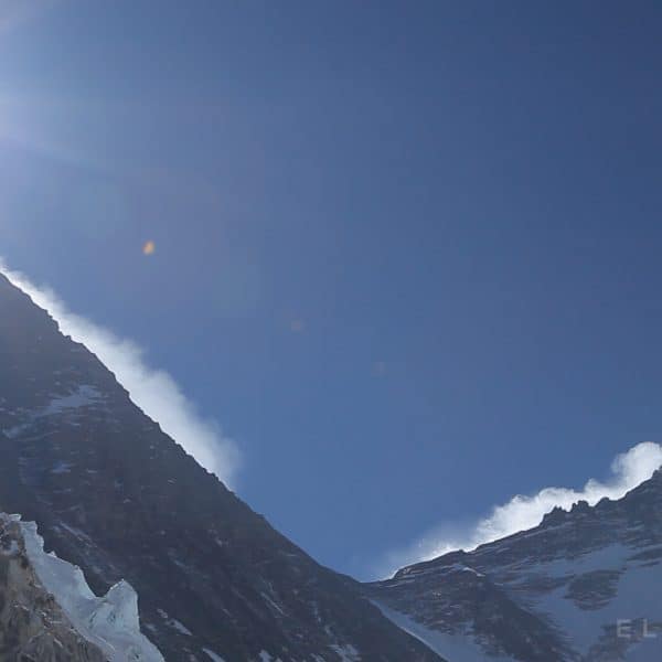 A tall dark mountain known as Everest stands next to another as clouds move across the peak with the bright sun shining high above