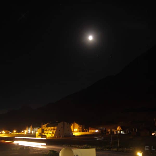 A small town lit by yellow lights in the Andes as the moon sets behind a mountains with a road and cars streaking past the camera