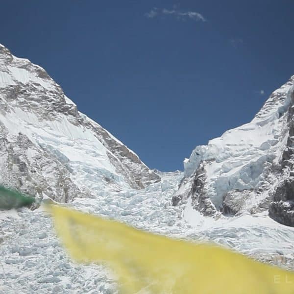 Yellow and green prayer flags in front of a large glacier called the Khumbu Icefall with two mountain peaks at the foot of Mt Everest