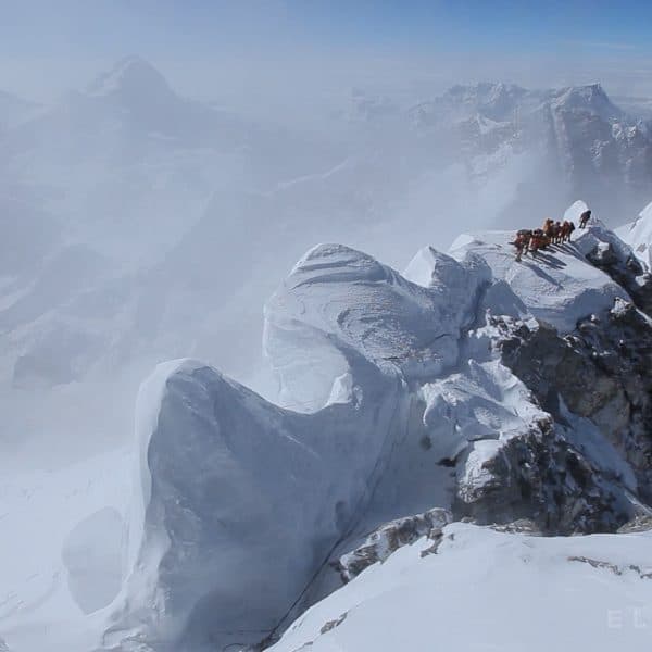 A snowy solid spire known as a cornice formed on top of a mountain next to a a team of climbers with the highest mountains in the world beneath them