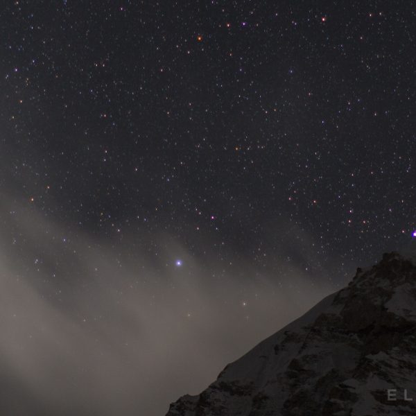 Close up a a Himalayan snow covered summit with clouds in the foreround and stars in the distance