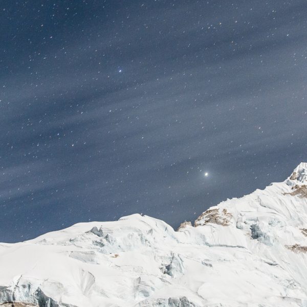 A triangular snow capped mountain peak with a vibrant blue night sky as stars appear to rise high above
