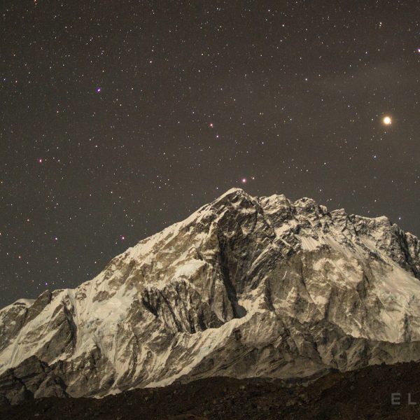 A large snow capped mountain in the Himalayas with stars rising at night near a rocky glacier
