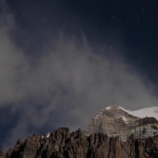 Summit of a rocky and snowy mountain at night with stars and cloud formations