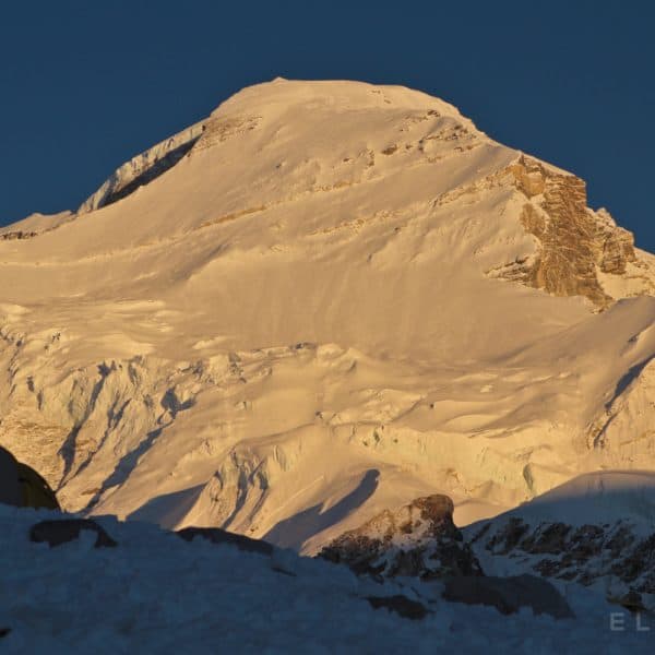 An impressive snow capped peak with yellow tents in the foregound with golden light illuminating the path to the summit
