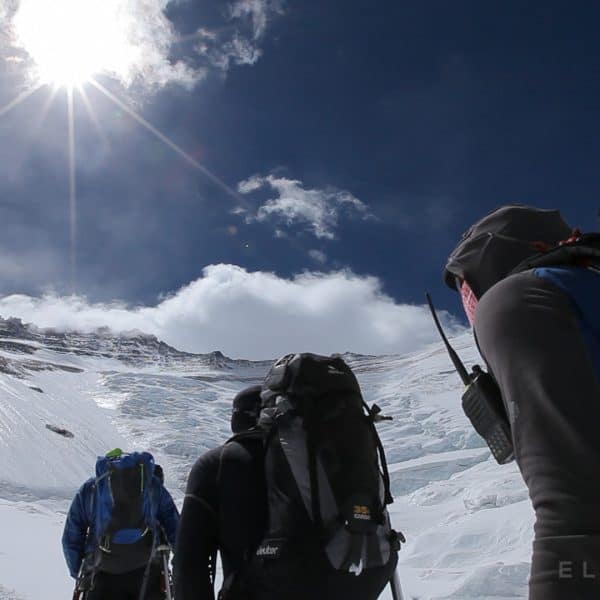 Climbers approach a steep icy vertical face with mountains in every direction as the sun rises high above