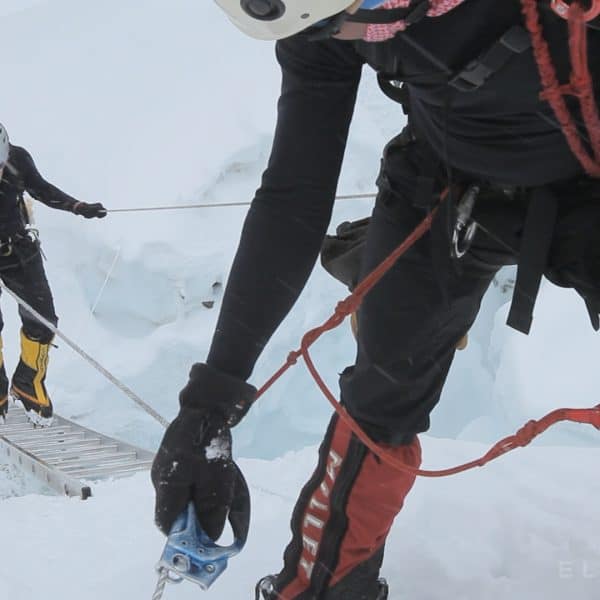 Two climbers assist another climber walk cross a horizontal ladder suspended over an open crevasse while holding the roppes