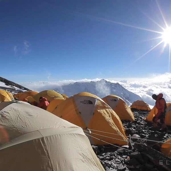 Yellow tents set up on a rocky glacier high above the clouds as climbers in multi colored down suits walk wowards a pile of oxygen cylinders