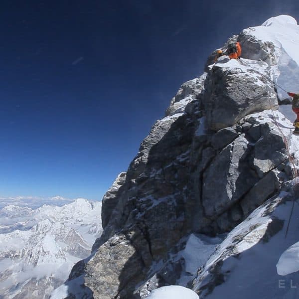Three climbers wearing multi colored warm down suits descend a stepp cliff called the Hillary Step on Mt Everest with mountains in the distance