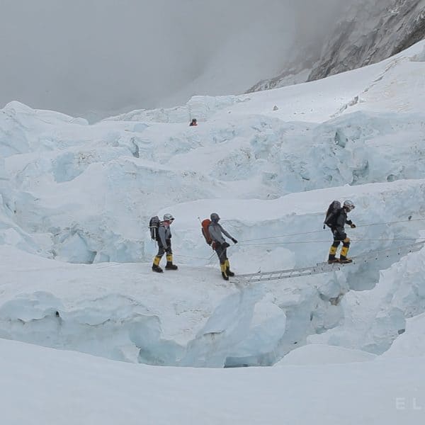3 climbers stand in a glaciated area on Mt Everest as one holds onto two ropes and walks across a dangerous ladder suspended over a crevassse