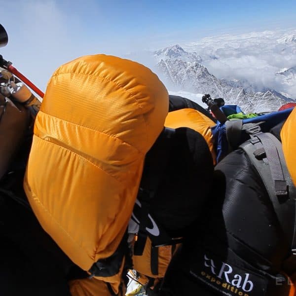 3 climbers in orange down suits hug on the summit of Everest with mountains in the distance while wearing oxygen masks and backpacks