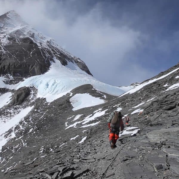 A climber wearing a red faded warm suit with a tall steep snow and rock mountain in the distance