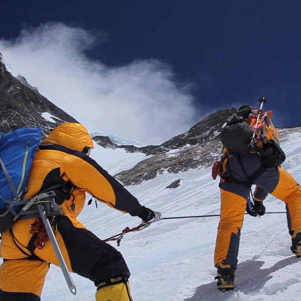 Two climbers walk in crampons dressed in warm yellow suits up a steep icy face with a tall mountain called Everest in the distance