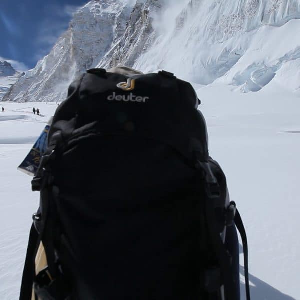 A climber walks on a snowy glacier carrying a backpack surrounded by mountains with 3 climbers in the distance