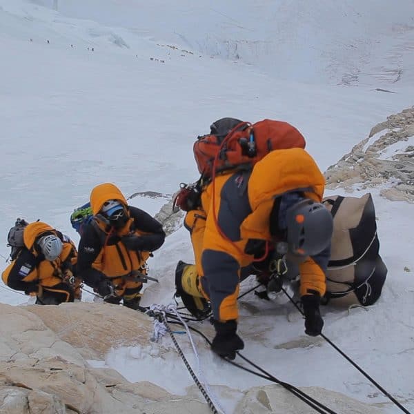 3 climbers ascend a yellow rock section on Mt Everest with a linek of climbers and yellow tents in the distance