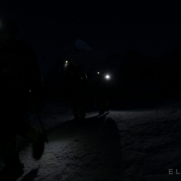 3 climbers walk in the dark with headlamps on a snowy glacier with a mountain in the distance