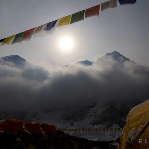 The moon rises above two mountains on a glacier with yellow tents and a sstring of prayer flags hangs in the foreground