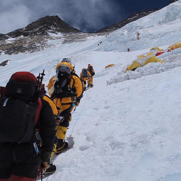 A small line up of climbers wearing yellow suits on a steep icy face with tents set up on exposed ledges with mountains high above